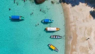 Top-down aerial view of a tropical bay with colorful tour boats anchored in crystal-clear turquoise water as people snorkel and swim near a sandy beach.