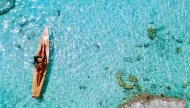 Top-down aerial view of a couple relaxing on a wooden canoe in crystal-clear, sparkling turquoise water on a sunny day.