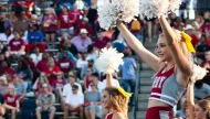 An enthusiastic Troy Trojans cheerleader with a yellow bow in her blonde hair, smiling and raising white pom-poms to energize the crowd at a football game.