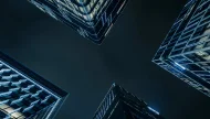 A dramatic low-angle, worm's-eye view looking up at the dark night sky framed by the corners of four modern skyscrapers with glowing blue and white architectural lights.