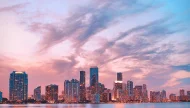 A beautiful panoramic view of the downtown Miami, Florida skyline with its illuminated skyscrapers reflecting on the water at sunset, under a dramatic pastel pink and blue cloudy sky.