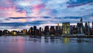 A dramatic sunset over the Midtown Manhattan skyline in New York City, with iconic buildings like the Empire State Building and Chrysler Building illuminated and reflecting on the East River.