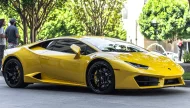 A striking yellow Lamborghini Huracán with black wheels parked on a city street on a sunny day, with lush green trees in the background.