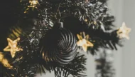 A moody, close-up photograph of a unique black spiral Christmas ornament hanging on a tree, with warm, glowing star-shaped fairy lights in the background.