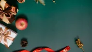 A festive Christmas flat lay featuring wrapped gifts, a red ribbon, pine cones, and gold decorations arranged as a border on a dark green background, leaving empty copy space in the center.