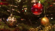 A warm, festive close-up of a Christmas tree decorated with a red snowflake ornament, reflective gold and silver baubles, and sparkling gold tinsel.