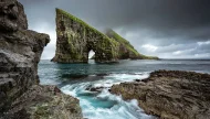 A dramatic long-exposure shot of the Drangarnir sea stacks and Tindhólmur islet in the Faroe Islands, with turbulent turquoise water swirling around the rocky coast under a stormy sky.