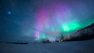 Vibrant green and purple Aurora Borealis (Northern Lights) dancing across a star-filled sky over a vast, snowy winter landscape.