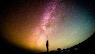Awe-inspiring silhouette of a person standing on a hill, looking up at the vibrant, colorful arc of the Milky Way galaxy in a vast, star-filled night sky.